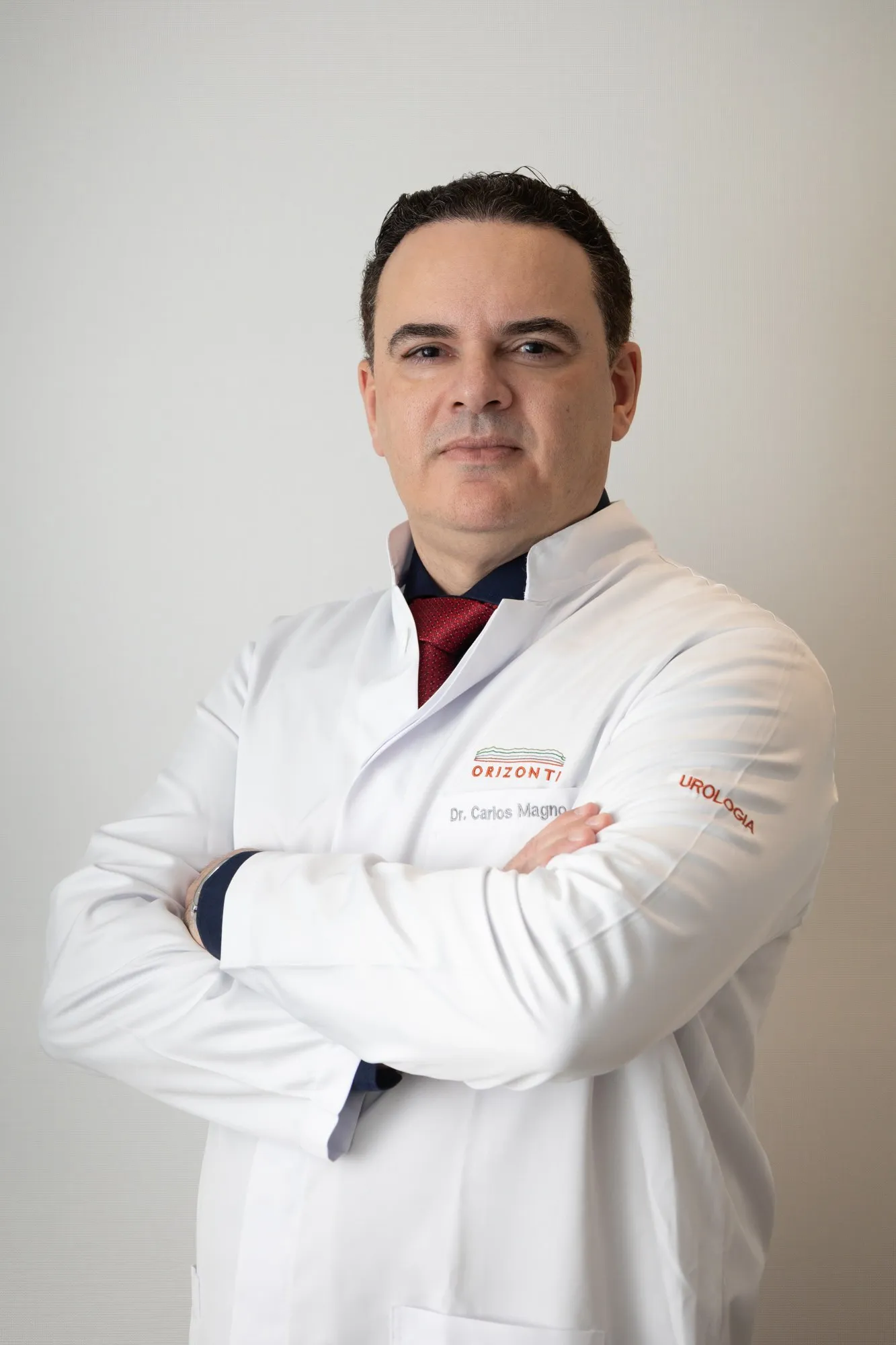 Dr. Carlos Magno de jaleco, braços cruzados, em fundo claro